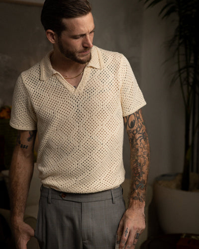 A man with tattoos on both arms wears the Dandy Del Mar Antibes Crochet Polo Shirt in Vintage Ivory and gray pants, standing indoors beside a potted plant.