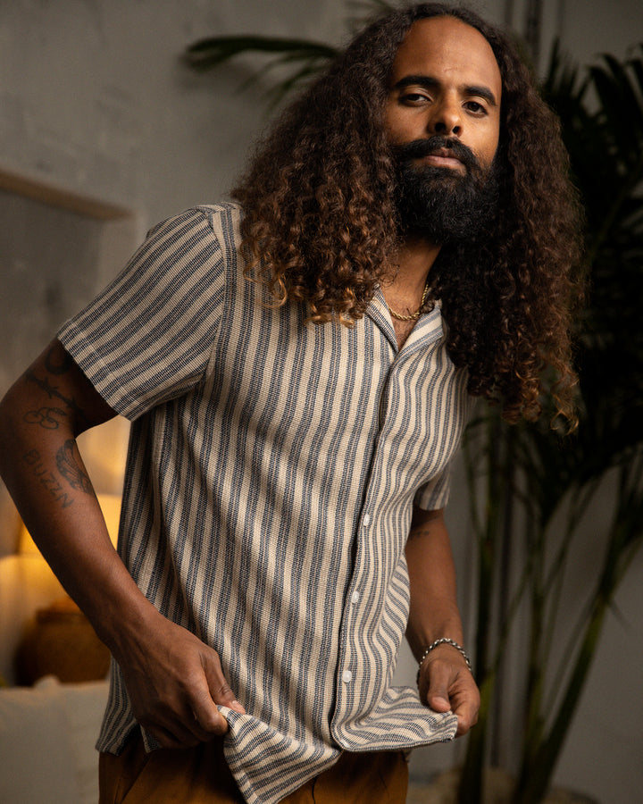 A man with long hair wearing a striped Dandy Del Mar Palma Shirt - Cacao Stripe.