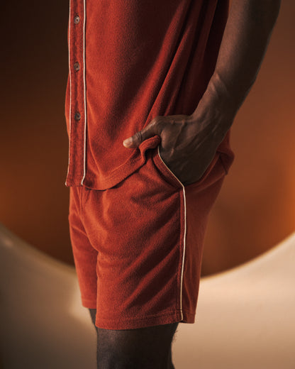 A person stands with one hand in their pocket, wearing Dandy Del Mar's "The Gaucho Short - Rouge," a rust-colored matching short-sleeve shirt and gaucho shorts with an elastic waistband, set against a gradient brown background.