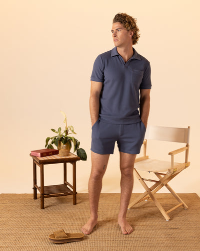 A man in the Dandy Del Mar Cannes Waffle Knit Polo Shirt in Moontide and shorts stands barefoot on a woven rug next to a chair, small table with books, potted plant, and one sandal.