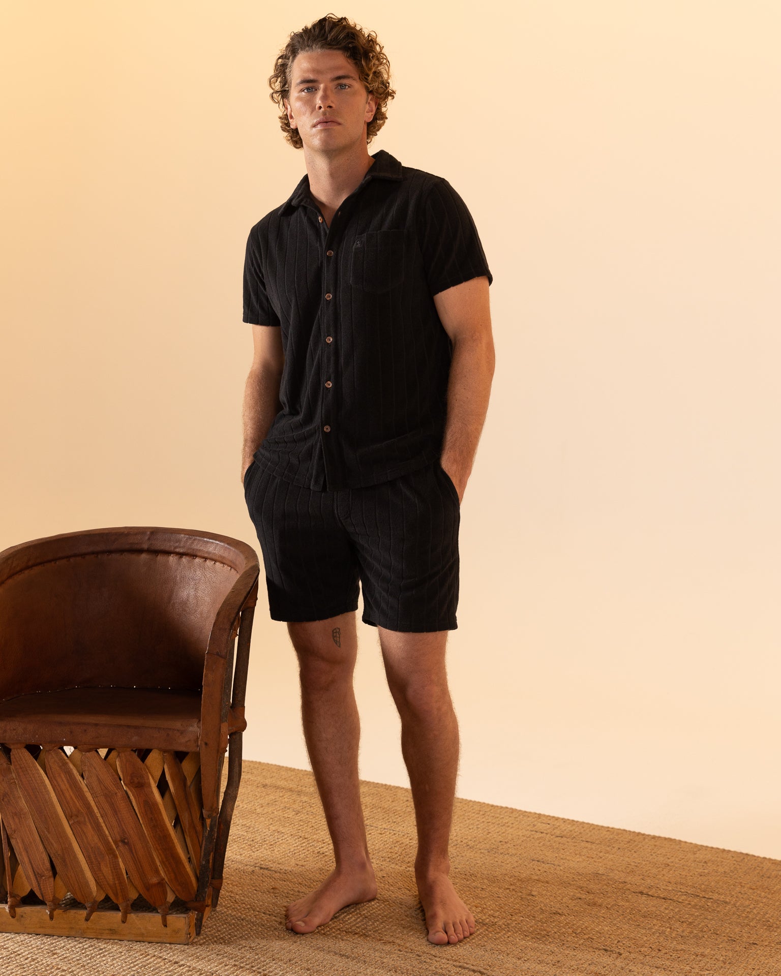 Barefoot and hands in pockets, a person stands next to a wooden chair on a woven rug against a beige backdrop, wearing the relaxed Côte d'Azur style of Dandy Del Mar's The Tropez Jacquard Shirt - Onyx with shorts.