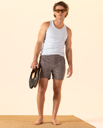 A man in a white tank top and The Corsica Corduroy Shorts - Argento by Dandy Del Mar stands barefoot on a mat, holding dark shoes in his left hand and wearing sunglasses against a plain background.