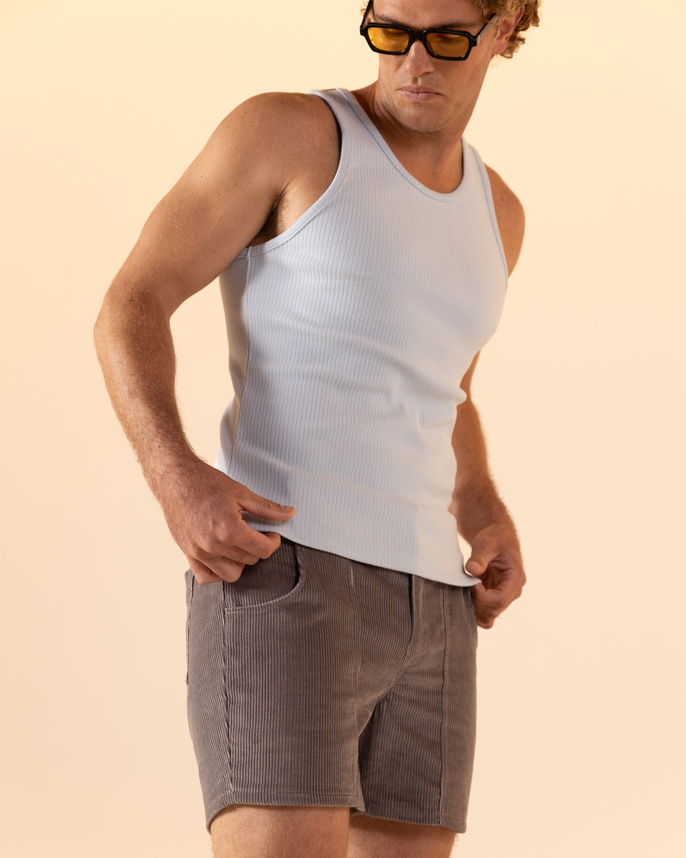 An individual in a white tank top, The Corsica Corduroy Short - Argento by Dandy Del Mar, and sunglasses gazes downward against a neutral background.