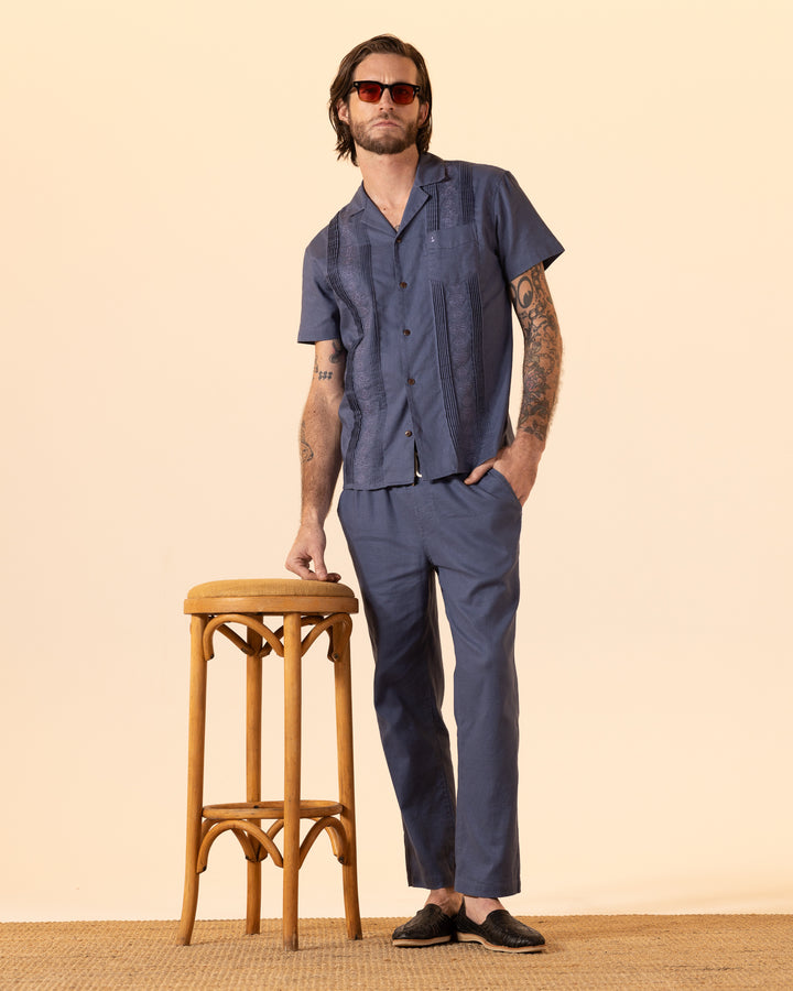 A man in sunglasses and a blue casual outfit, wearing The Brisa Linen Pant - Moontide by Dandy Del Mar, stands next to a wooden stool.