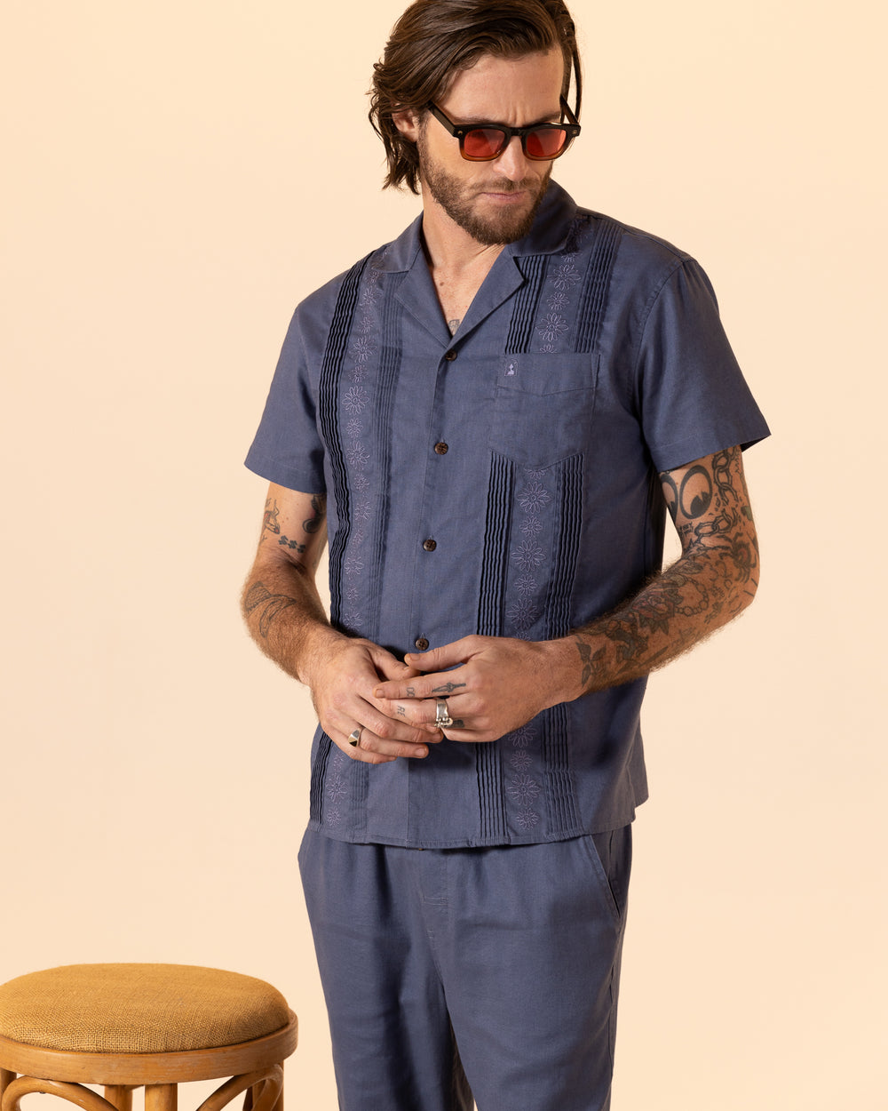 A bearded man wearing sunglasses and The Brisa Linen Shirt - Moontide from Dandy Del Mar stands next to a wooden stool with a beige cushion.