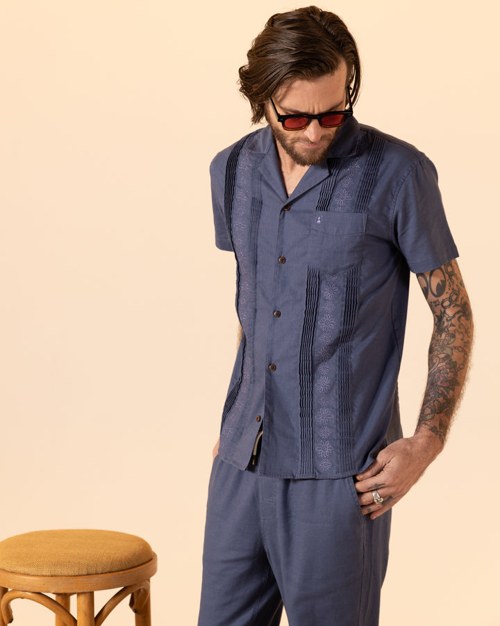 A man in sunglasses wears a blue short-sleeve shirt and Dandy Del Mar's The Brisa Linen Pant in Moontide, standing by a stool.