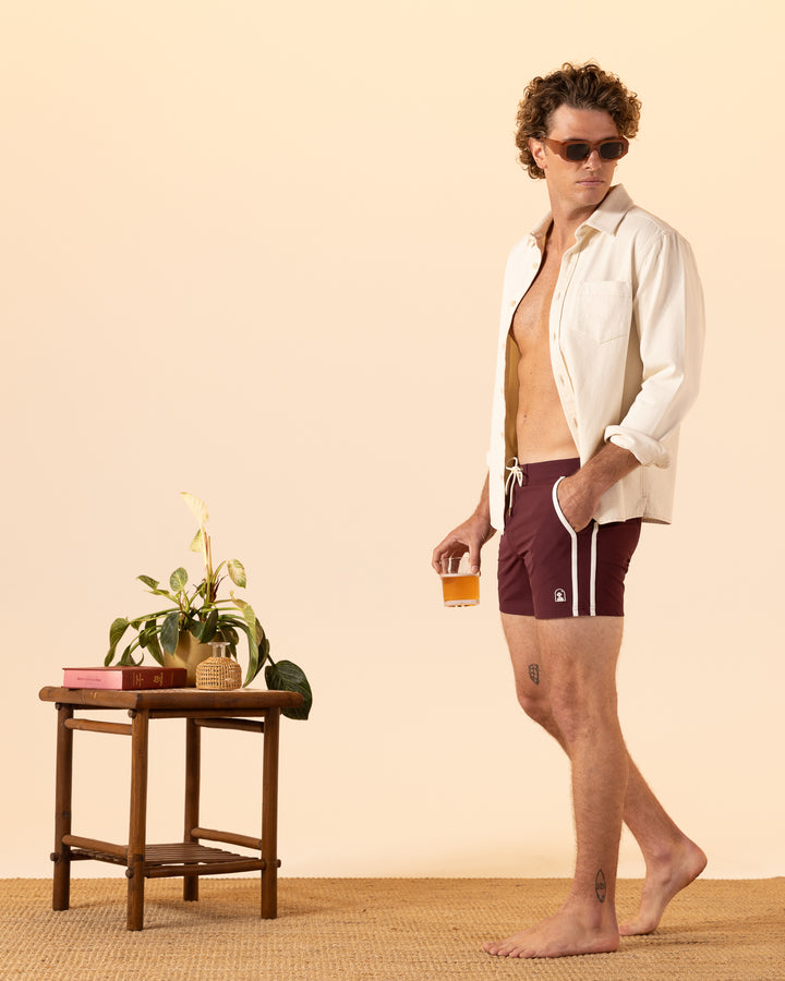 A man wearing The Stirata Swim Short - Bordeaux from Dandy Del Mar stands holding a drink near a small table with a plant and books.