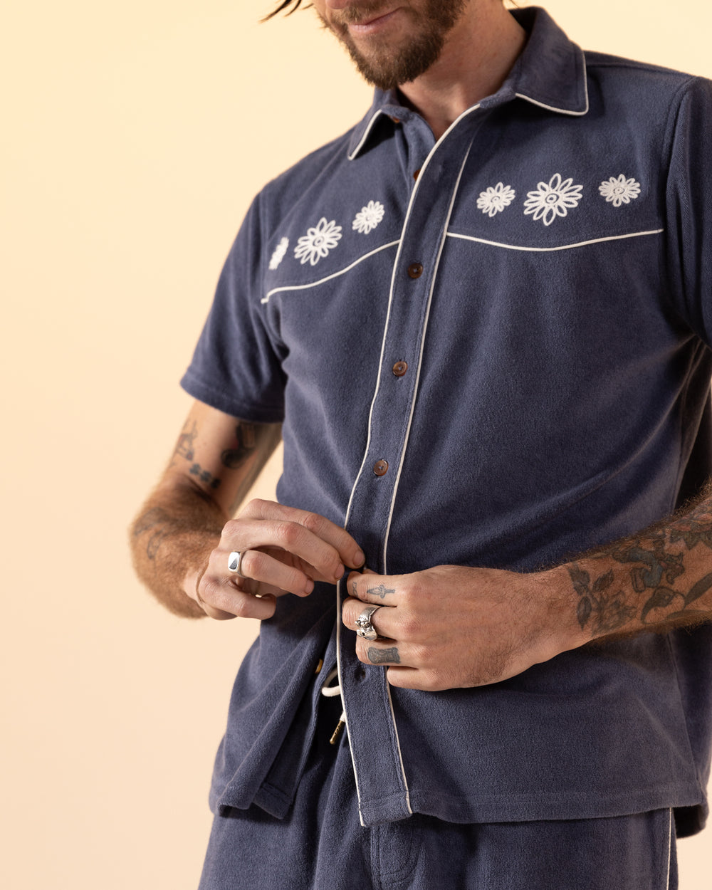 A person adorned with tattoos and rings fastens a button on The Gaucho Terry Cloth Shirt - Moontide by Dandy Del Mar, a blue short-sleeve shirt featuring floral embroidery paired with matching pants.