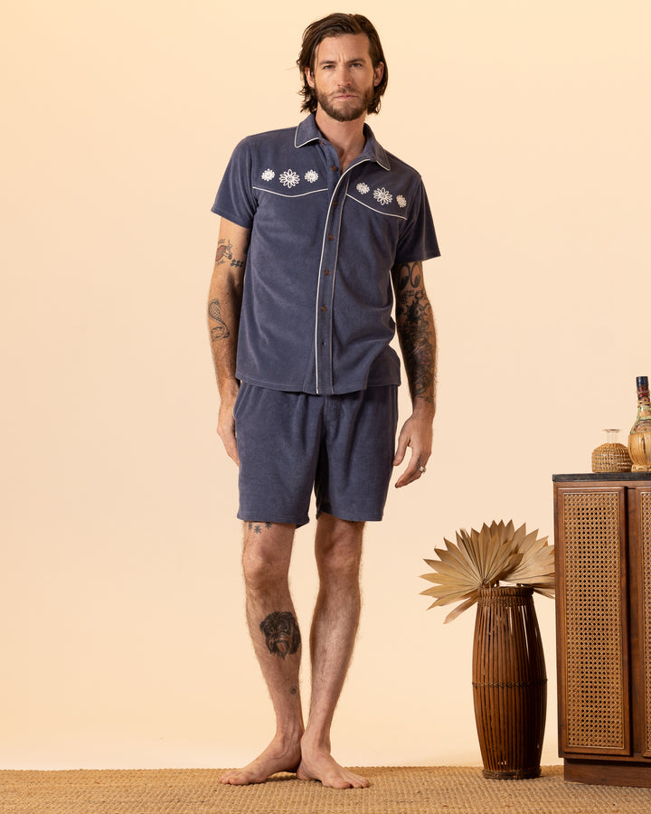 A tattooed individual stands barefoot on a woven mat, dressed in a blue The Gaucho Terry Cloth Shirt - Moontide and shorts from Dandy Del Mar. They are beside a wooden cabinet adorned with decorative items and a vase filled with dried leaves.