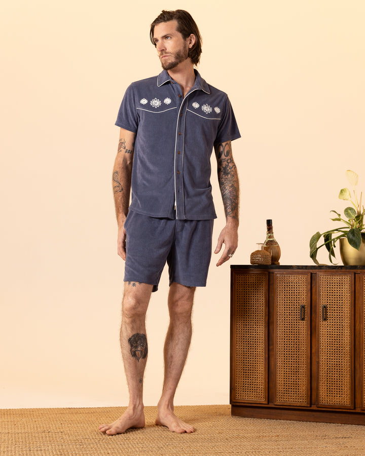 A man in a blue embroidered shirt and Dandy Del Mar's Gaucho Terry Cloth Shorts in Moontide stands barefoot by a wooden cabinet with plants.