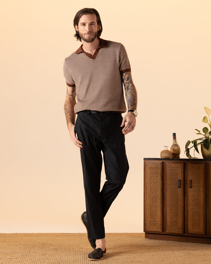 A tattooed man stands casually wearing The Palamos Knit Polo Shirt in Cappuccino by Dandy Del Mar, paired with dark pants. He is beside a mid-century modern cabinet with a plant and decorative bottles on top.