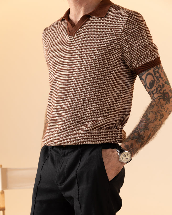 Person sporting The Palamos Knit Polo Shirt in Cappuccino by Dandy Del Mar and black pants, with hands in pockets. Visible arm tattoos and a watch complete the look.