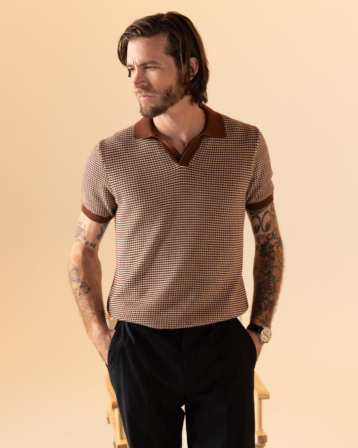 A tattooed person is wearing The Palamos Knit Polo Shirt - Cappuccino by Dandy Del Mar with dark pants, standing against a plain beige background, glancing sideways.