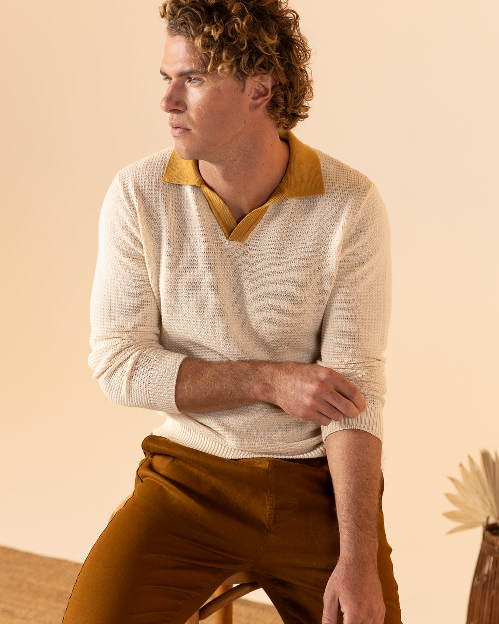 A person with curly hair sits on a stool, wearing The Sebastian Long Sleeve Knit Polo Shirt in Vintage Ivory by Dandy Del Mar and brown pants, gazing to the left.