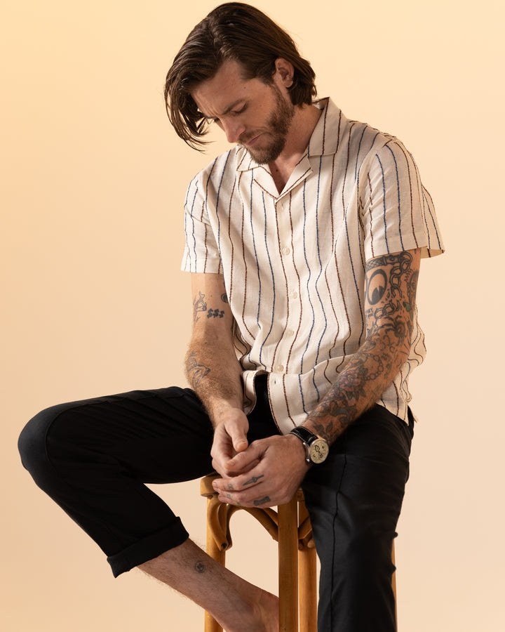 A tattooed man in Dandy Del Mar's The Quarta Shirt - Scallop Weave and dark pants sits on a stool, gazing downward against a beige backdrop.