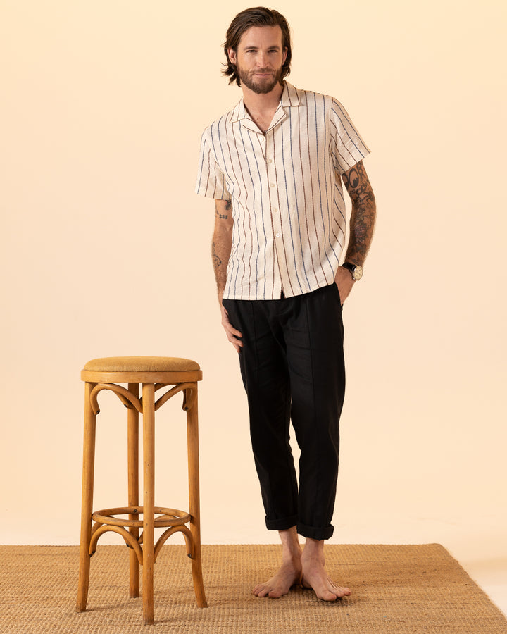 A man with a beard stands barefoot on a woven mat near a wooden stool, wearing The Quarta Shirt - Scallop Weave by Dandy Del Mar and dark pants. He smiles with his hands in his pockets against a beige background.