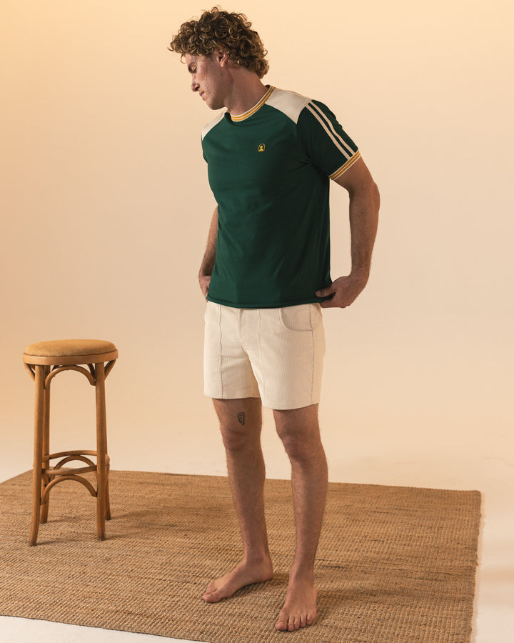 A man stands barefoot on a woven rug in cream shorts and The Nassau Tee - Albero by Dandy Del Mar, beside a wooden stool, against a neutral background.