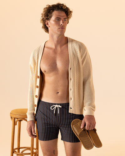 A man in a cream open-knit cardigan and Dandy Del Mar’s The Ventura Volley Shorts - Moontide, featuring an elastic waist, stands holding sandals beside a wooden stool against a beige background.