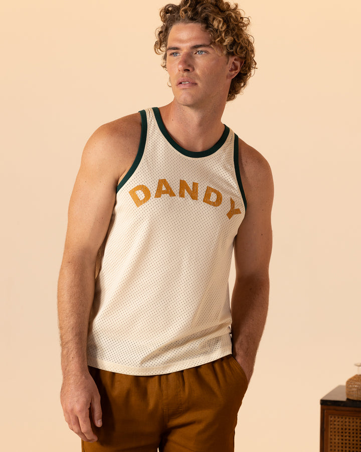 A curly-haired man stands against a beige background wearing The Kaena Mesh Tank - Sabbia by Dandy Del Mar.