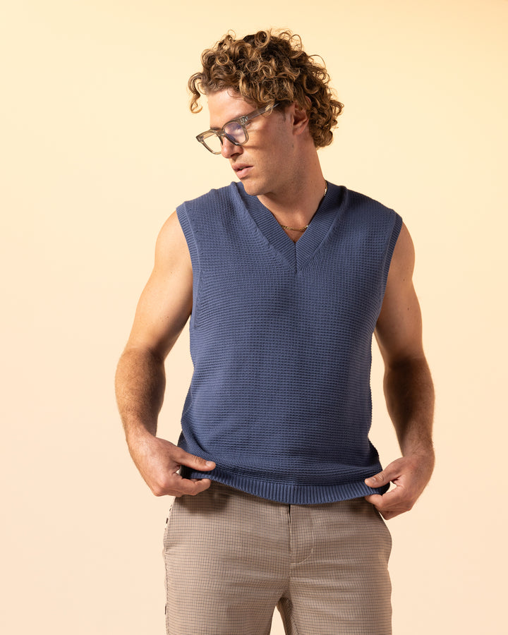 A person with curly hair and glasses is wearing The Montego Knit Vest - Moontide by Dandy Del Mar and beige pants, looking down against a light beige background.