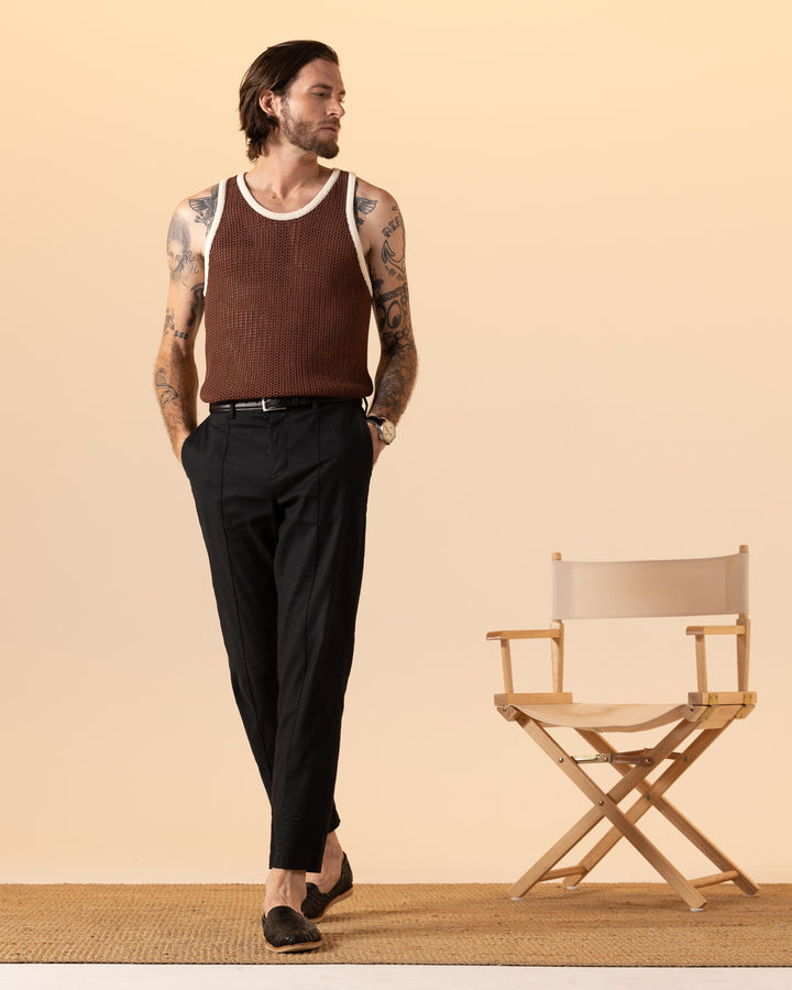 A person stands on a rug, wearing The Dominica Crochet Tank in cappuccino by Dandy Del Mar and black pants, next to an empty director's chair against a beige background.