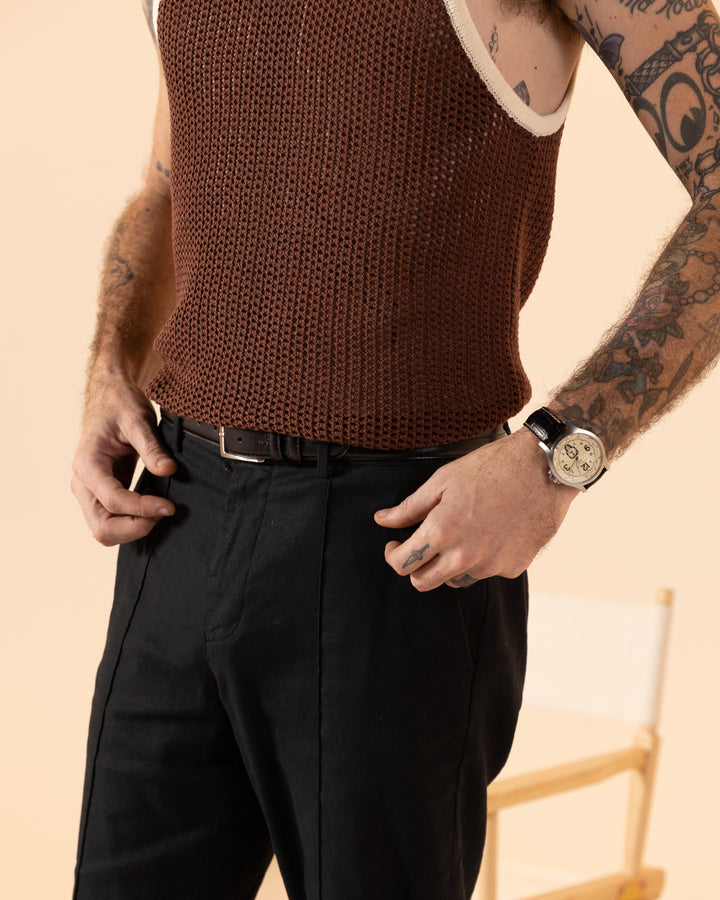 A tattooed individual stands near a beige chair, effortlessly wearing The Dominica Crochet Tank in cappuccino by Dandy Del Mar and black pants, paired with a watch.