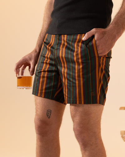 A person wearing Dandy Del Mar’s The Mallorca Swim-Walk Shorts in Burnt Sienna and a black shirt holds a glass of amber liquid, with one hand in their pocket and a tattoo visible on their knee.