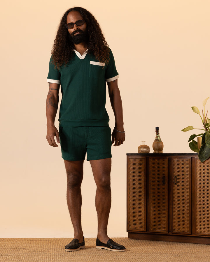 A person with long curly hair and a beard stands in The Cannes Waffle Knit Shorts - Albero by Dandy Del Mar, wearing sunglasses next to a wooden sideboard with plants and a bottle.