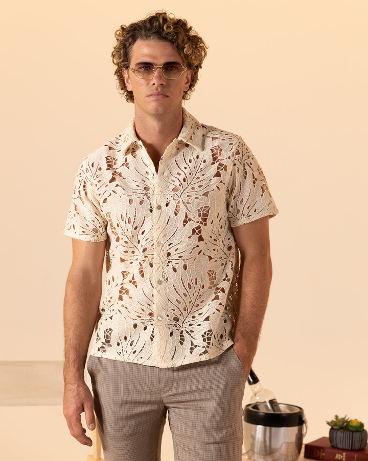 A man with curly hair wearing sunglasses and The Monstera Shirt - Scallop by Dandy Del Mar stands in front of a neutral background, with a chair, plant, and ice bucket partially visible beside him.