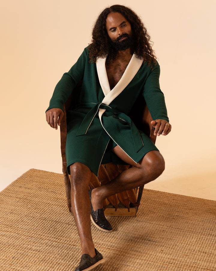 A person with long curly hair sits on a chair wearing the Cannes Waffle Knit Robe - Albero from Dandy Del Mar paired with black slip-on shoes against a beige backdrop.