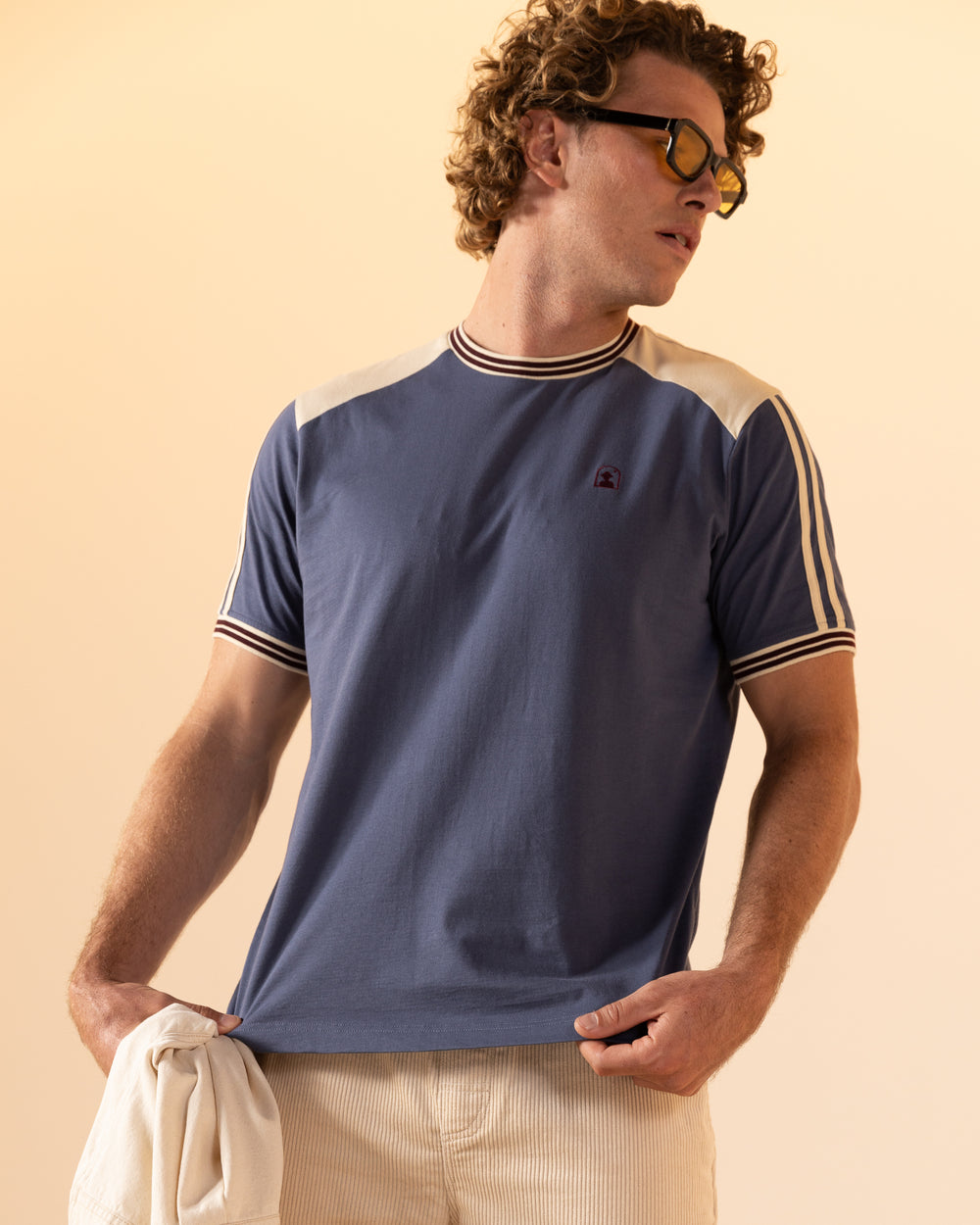 A person in a blue and cream The Nassau Tee - Moontide by Dandy Del Mar, featuring an embroidered logo, holds a light jacket and wears sunglasses against a plain background.