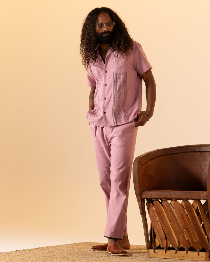 A long-haired, bearded person wears glasses and The Brisa Linen Shirt - Dawn by Dandy Del Mar, along with matching pants. They stand on a rug next to a wooden chair against a beige backdrop.