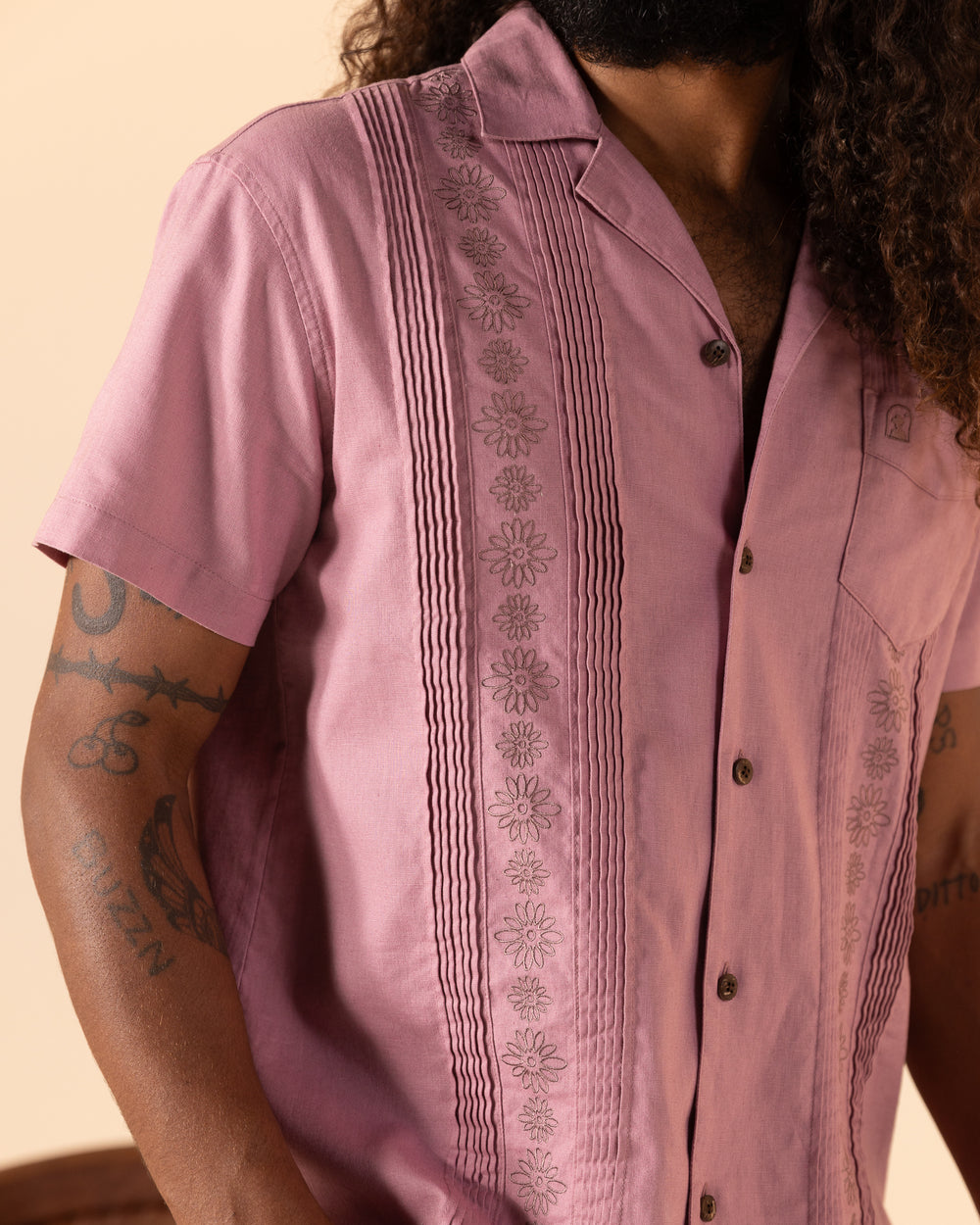 A person wearing The Brisa Linen Shirt - Dawn from Dandy Del Mar, featuring pink floral embroidery and short sleeves, reveals tattoos on their arms.