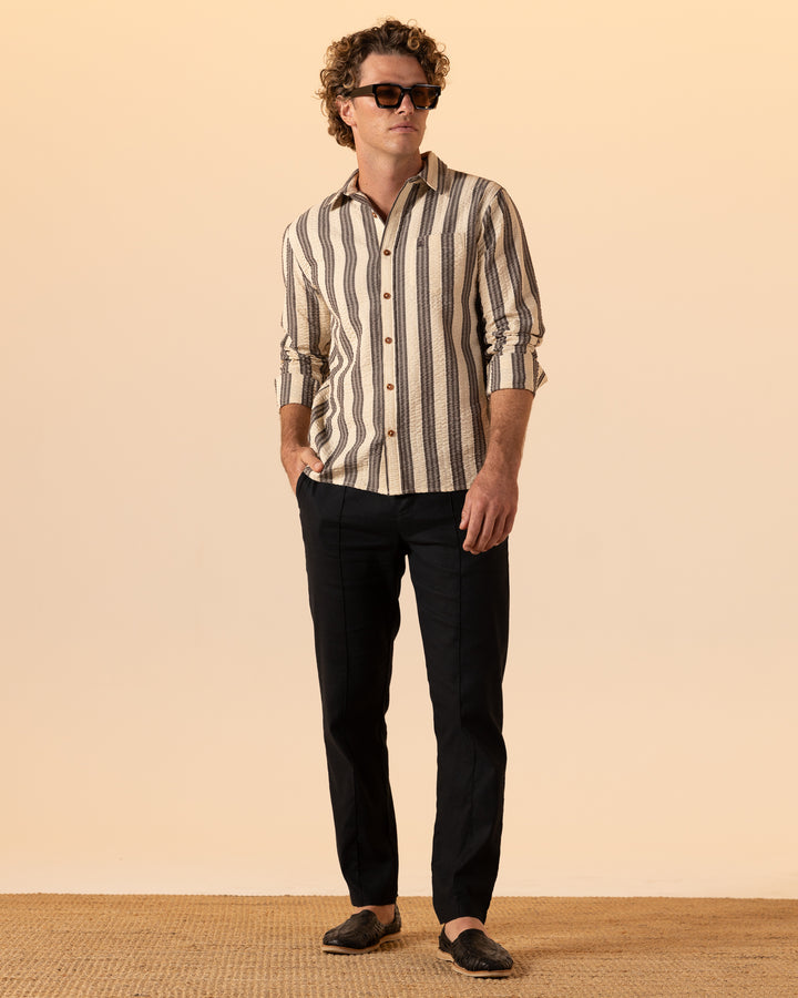 A man wearing The Santos Shirt - Tierra Weave by Dandy Del Mar, dark trousers, brown shoes, and sunglasses stands on a textured beige floor against a light beige background.