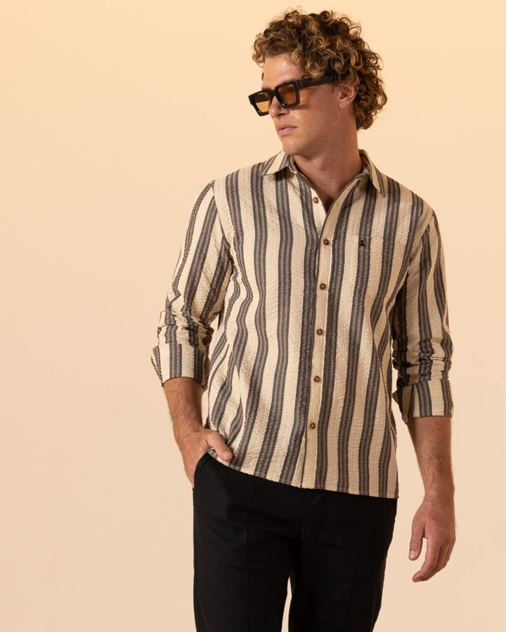 Someone wearing sunglasses and The Santos Shirt - Tierra Weave by Dandy Del Mar, paired with black pants, stands on a woven rug against a plain background.