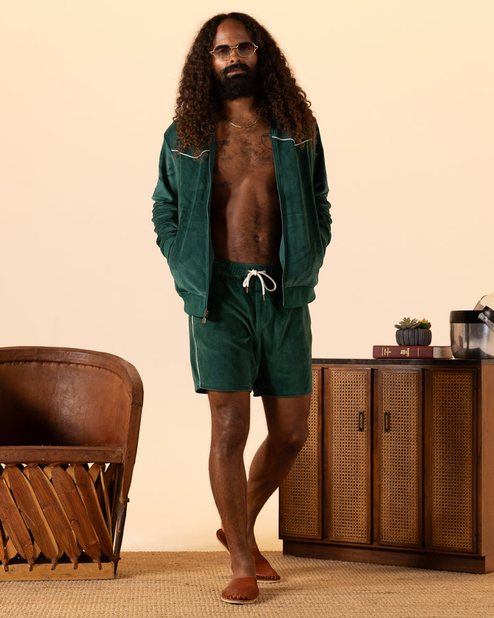 A man with long curly hair and a beard wears The Rio Velour Short - Albero by Dandy Del Mar, paired with sandals and glasses. He stands beside a wooden chair and cabinet against a neutral backdrop.