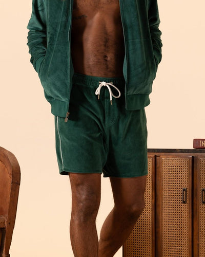 A person stands indoors with hands in pockets, wearing a dark green zip-up jacket and The Rio Velour Shorts - Albero by Dandy Del Mar, featuring an elastic waistband.