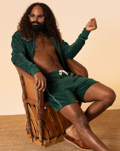 A man with long curly hair and a beard sits on a wooden chair, wearing sunglasses, a green velour jacket, Dandy Del Mar's The Rio Velour Shorts - Albero, and brown sandals on a beige woven rug.