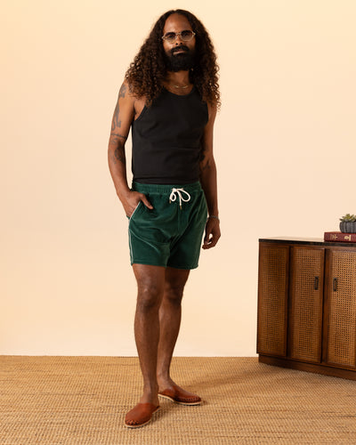 A man with long curly hair, in a black tank top and Dandy Del Mar’s The Rio Velour Shorts - Albero featuring an elastic waistband, stands on a woven rug beside a wooden cabinet with books and a small plant.