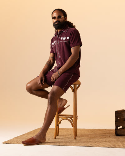 Sporting a full beard and long hair, a man wearing The Gaucho Short in Bordeaux by Dandy Del Mar sits on a wooden stool atop a woven rug, perfectly embodying the Equatorial Cowboy look.