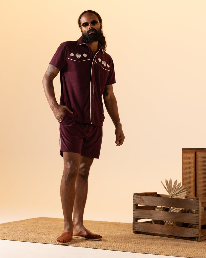 A person wearing Dandy Del Mar's The Gaucho Terry Cloth Short in Bordeaux stands on a woven rug beside a wooden crate with decorative objects, set against a beige background.