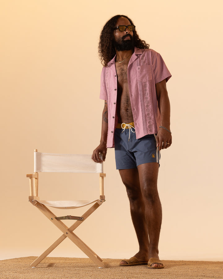 A person with long hair and sunglasses stands beside a director's chair, wearing a pink shirt, Dandy Del Mar's Stirata Swim Shorts in Moontide, and sandals.