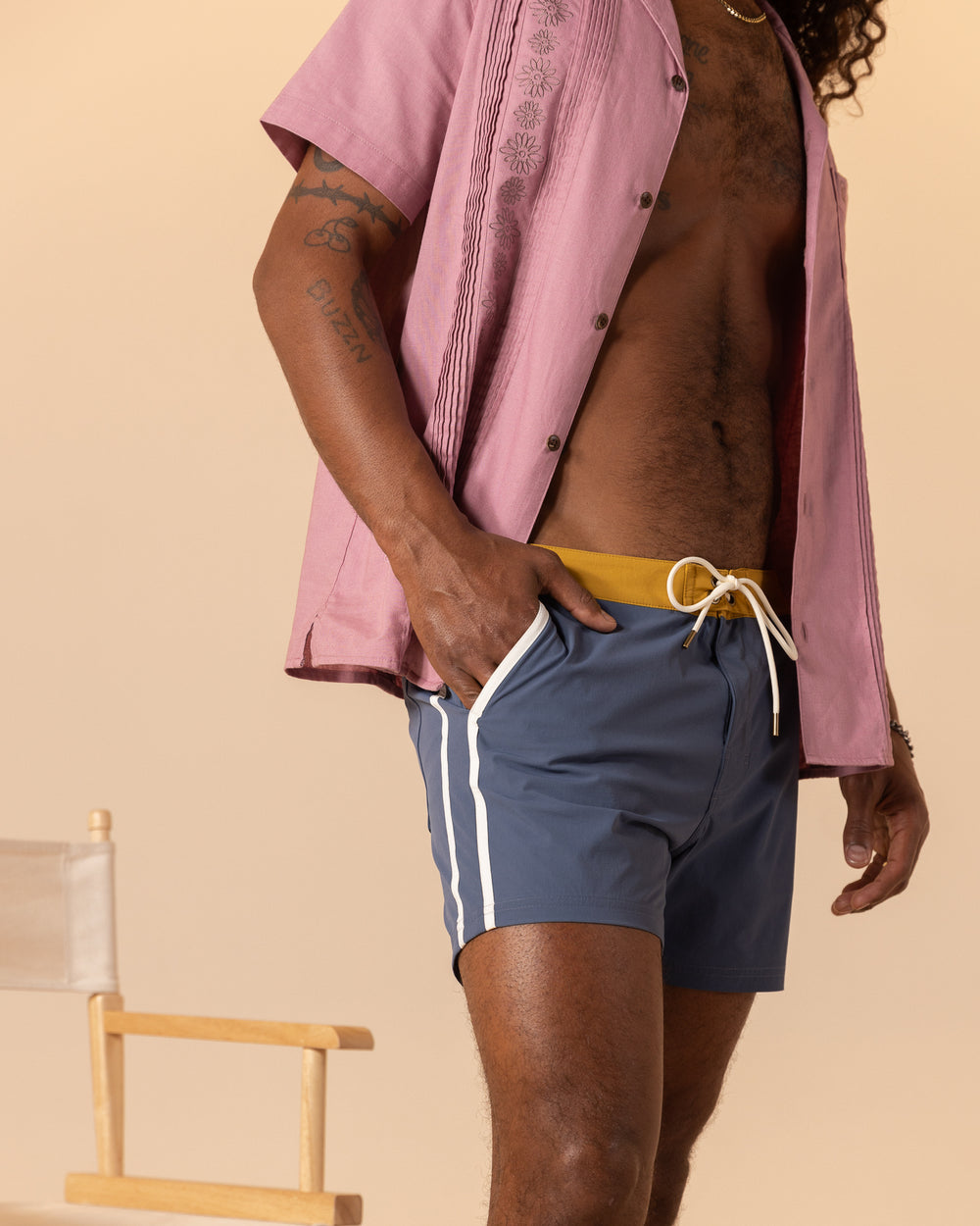 A person wearing a pink short-sleeve shirt and the Moontide Stirata Swim Short by Dandy Del Mar stands by a wooden chair against a beige background.