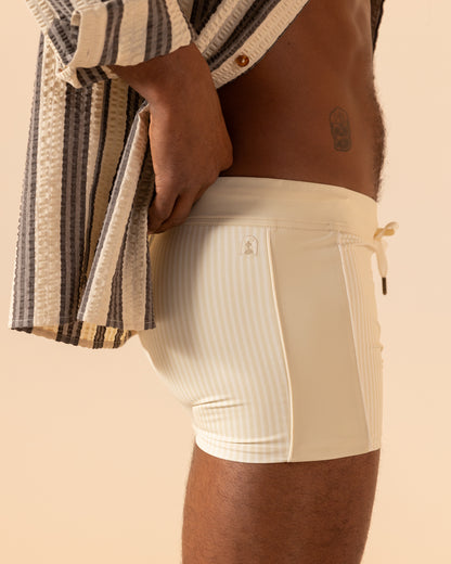 A person wearing Dandy Del Mar's The Cassis Square Cut Swim Brief - Sabbia stands in profile against a beige background, exuding a lust for leisure. This ensemble adds an effortlessly stylish touch to their relaxed demeanor.