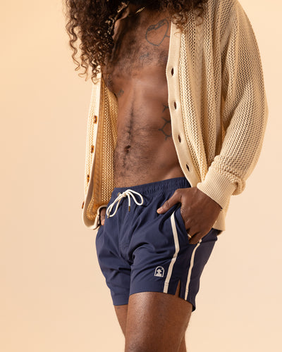 A person wears an open beige cardigan and Dandy Del Mar's The Ventura Volley Shorts - Anchor, revealing part of their chest and midsection against a plain background.