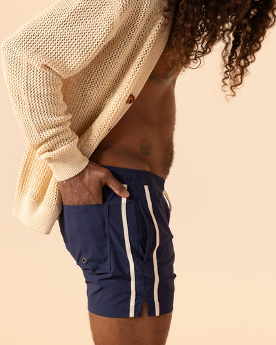 A person with long curly hair stands sideways with hands in pockets, wearing a cream knit cardigan and the Ventura Volley Shorts - Anchor by Dandy Del Mar.