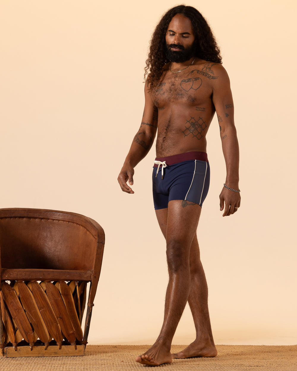 A person with long curly hair and tattoos is wearing The Cassis Square-Cut Swim Brief - Anchor by Dandy Del Mar, standing next to a brown chair on a beige background.