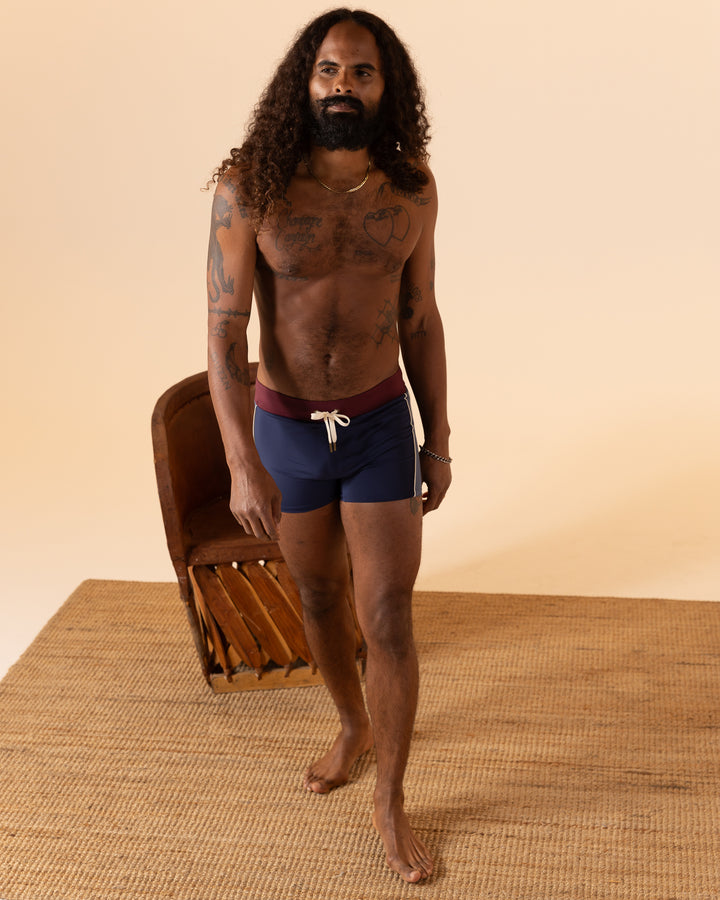 A person with long curly hair and tattoos is walking on a woven mat wearing The Cassis Square-Cut Swim Brief by Dandy Del Mar, with a chair in the background.