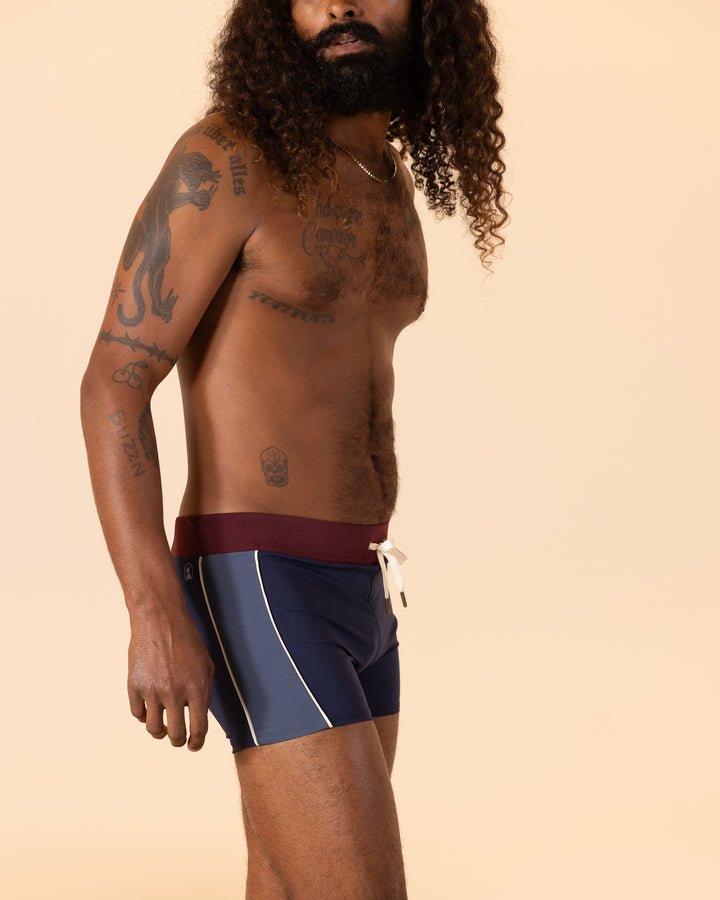 A person with long curly hair and tattoos wears The Cassis Square-Cut Swim Brief - Anchor by Dandy Del Mar, standing against a plain beige background.