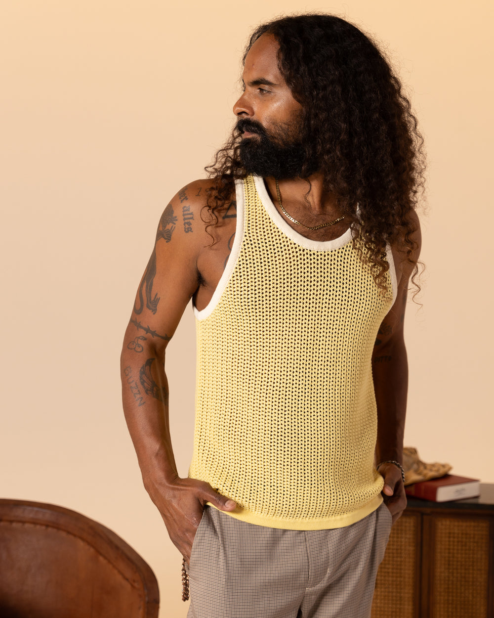 A person with long curly hair and tattoos wears The Dominica Crochet Tank in Plantain by Dandy Del Mar, paired with checkered pants, standing against a neutral background.